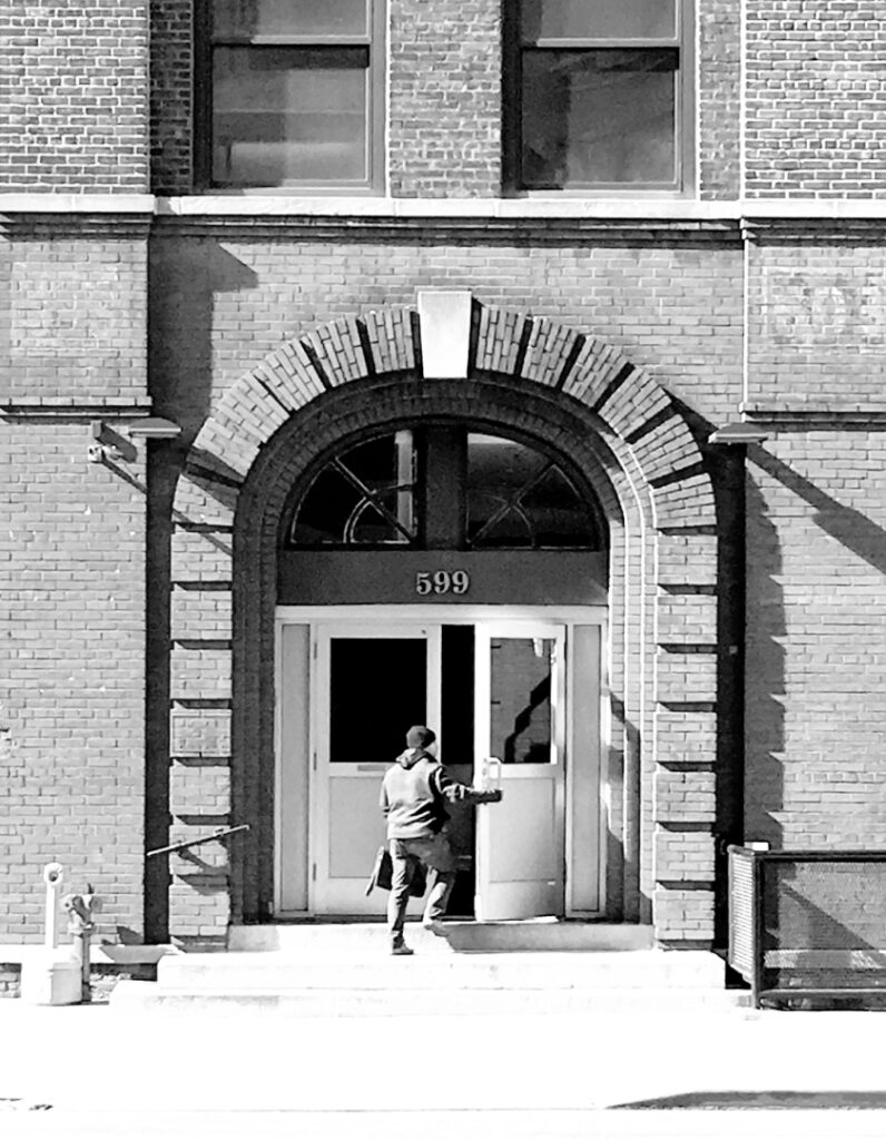 A black and white photo of a man walking through the front door of a large brick building.